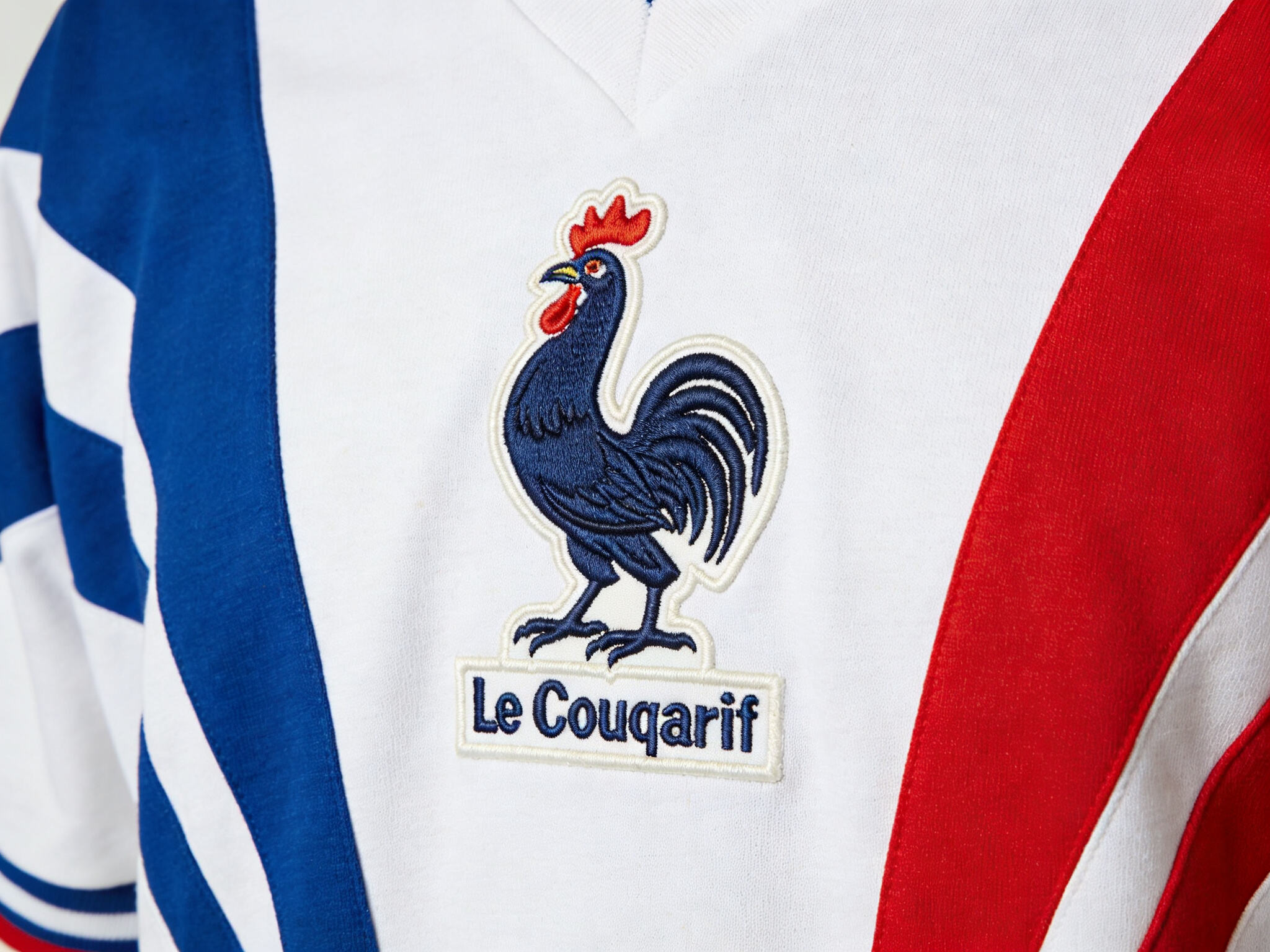 A detailed close-up of the classic France 1984 home jersey by Le Coq Sportif, highlighting the large, embroidered rooster crest on the chest and the distinctive blue, white, and red tricolor stripes running down the sides.